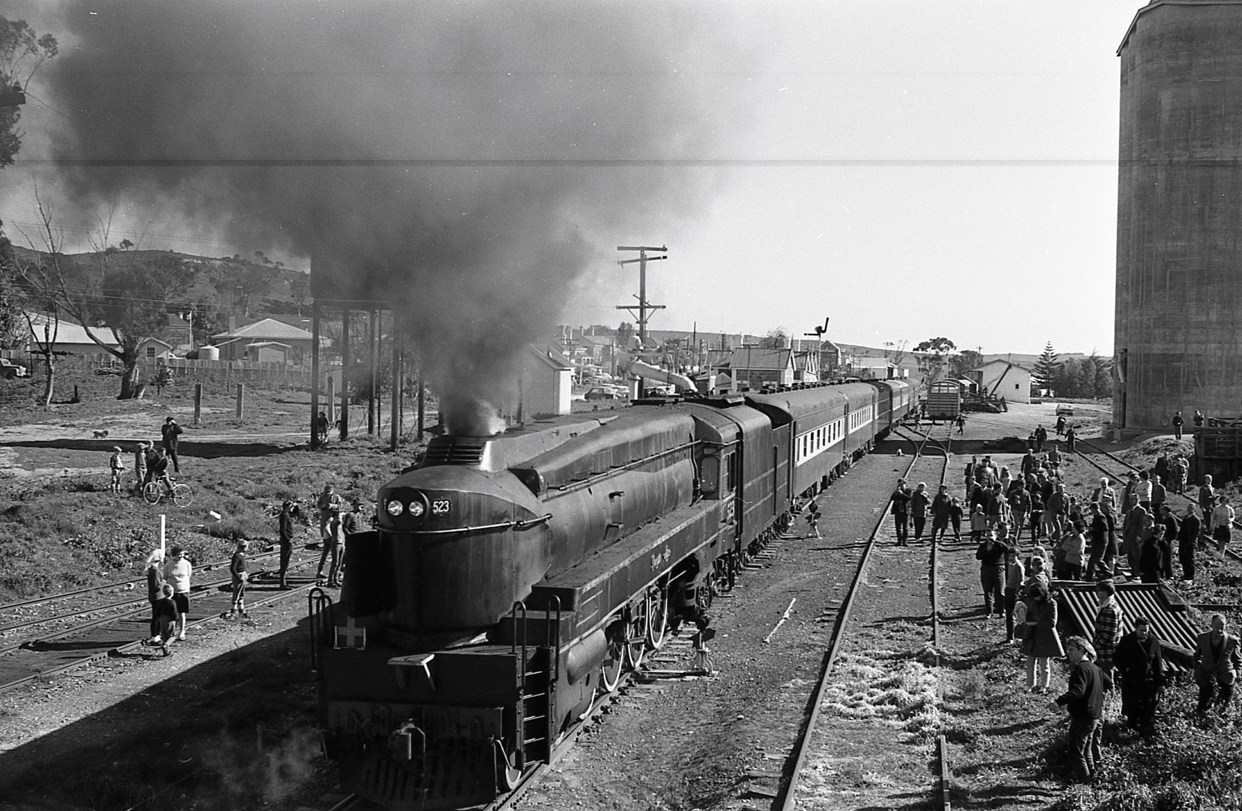 Black and white historic photo at Eudunda in 1960's of a large steam locomotive running along train tracks with a crowd of people at a rustic t...